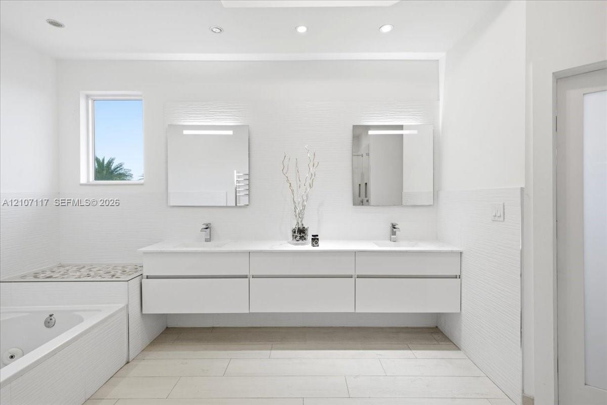 Floating white double vanity with polished stone countertop, rectangular undermount sinks, wall-mounted light fixtures, tiled bathtub surround with stone inlay, and tile flooring