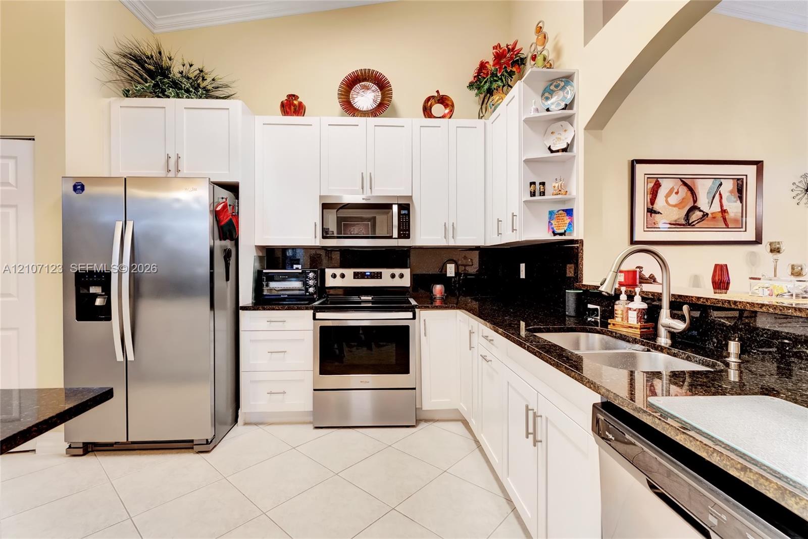 Kitchen featuring white shaker-style cabinetry, dark stone countertops, stainless steel appliances, a dual-basin undermount sink with a gooseneck faucet, and light-toned tile flooring