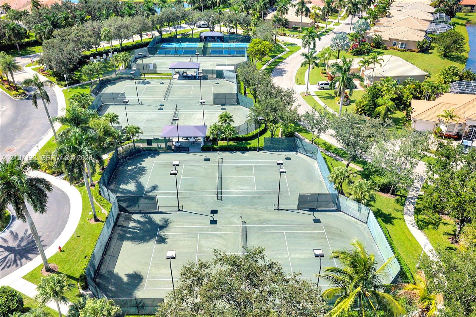Multi-court tennis facility featuring hard-surface playing areas, fenced perimeters, overhead lighting, and covered spectator seating