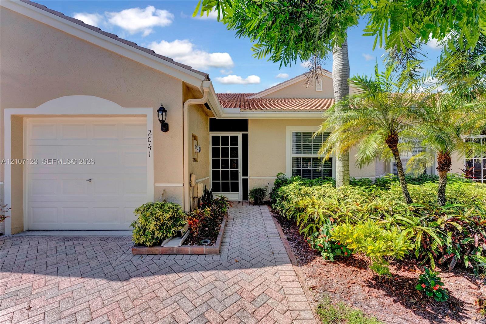 Single-story exterior featuring a clay tile roof, stucco facade, paver driveway, walkway, and glass-paneled front door