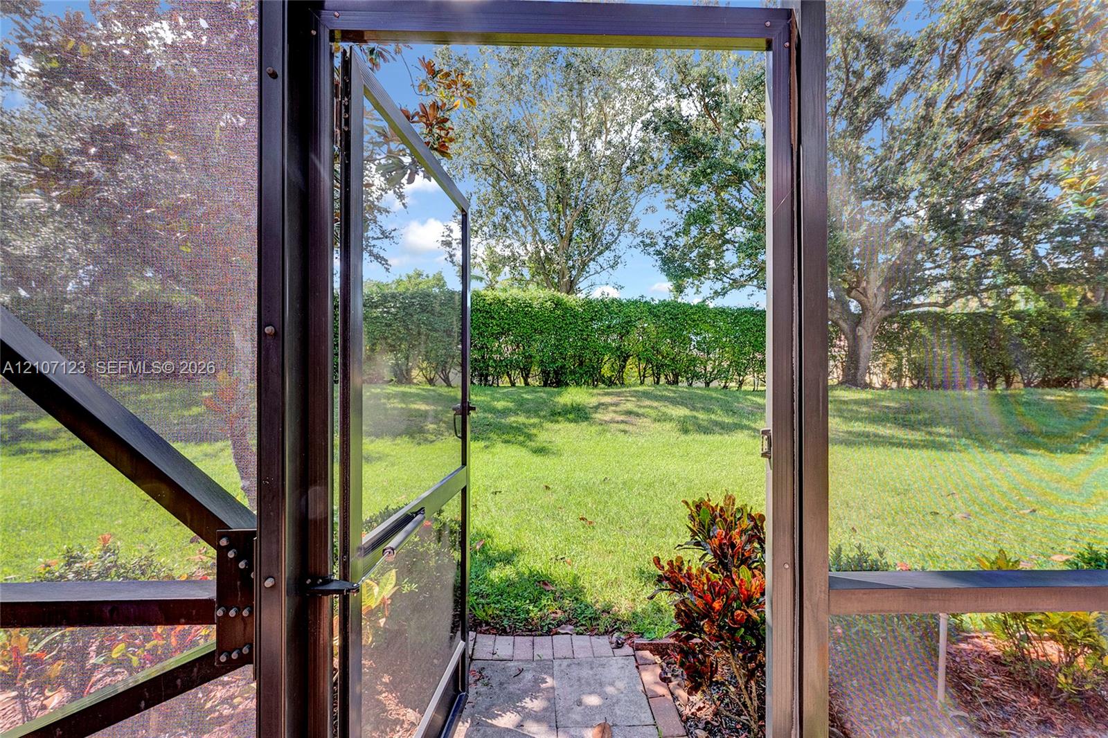Screened enclosure featuring a metal-frame door, stone paver threshold, mature trees, manicured lawn, and natural hedge perimeter