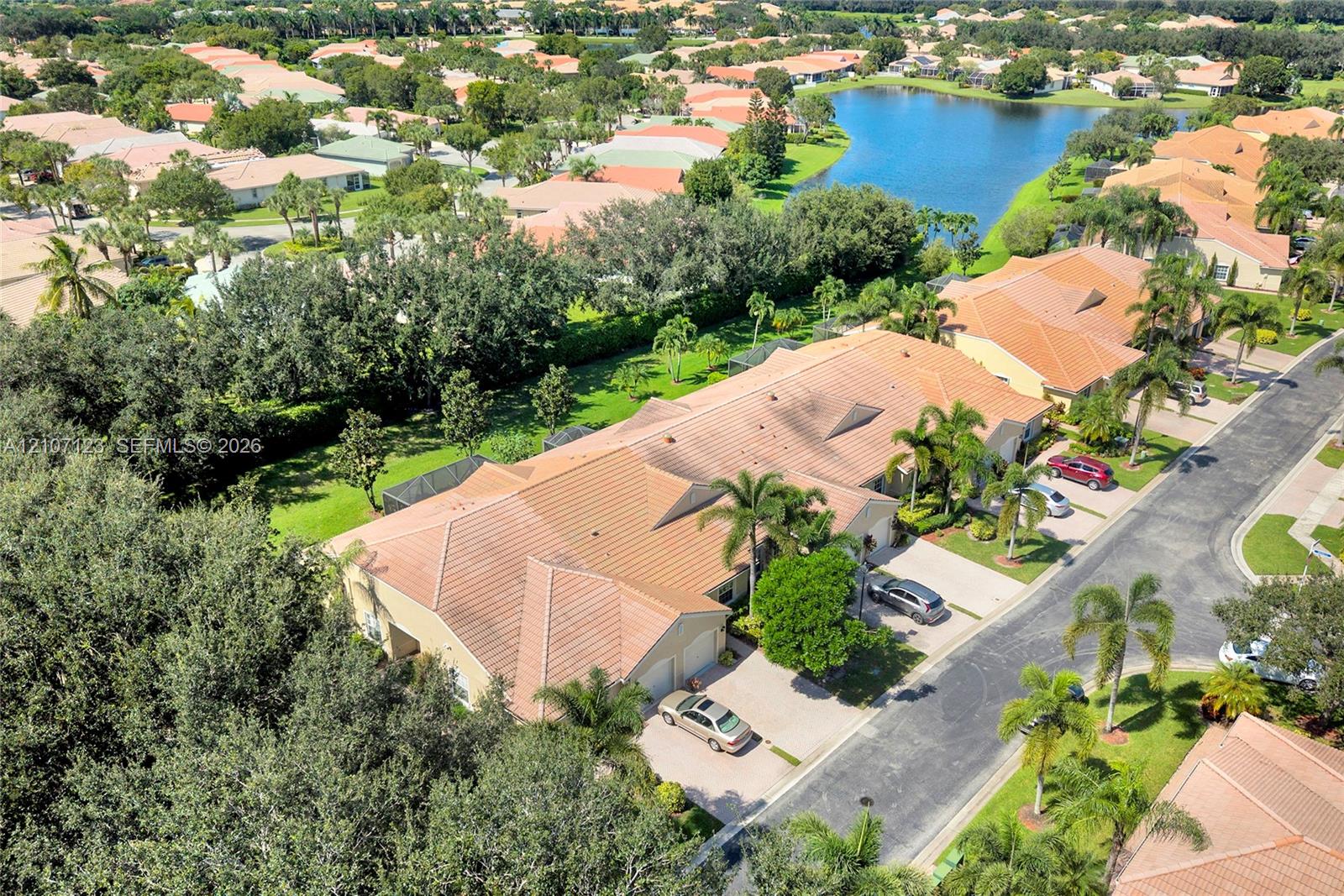 Aerial perspective featuring a tile roof, paved driveway, mature trees, and proximity to a community lake