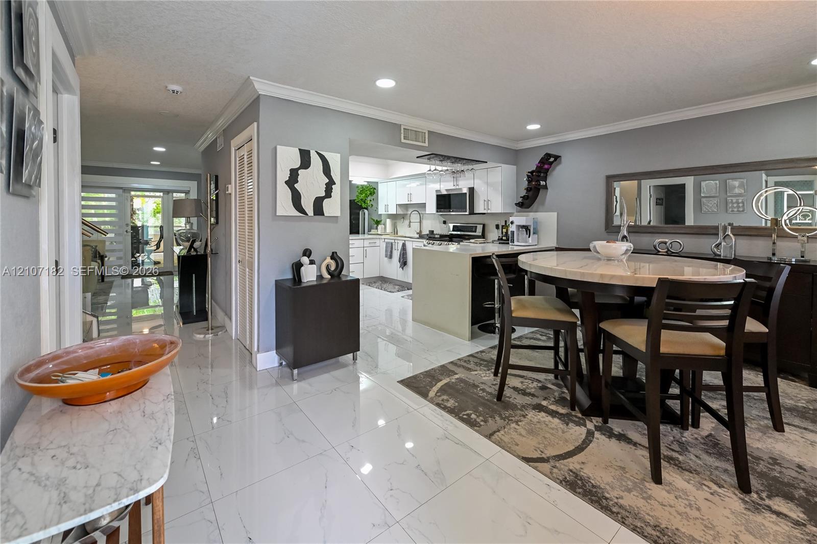 Open-concept interior featuring large-format polished tile flooring, recessed lighting, crown molding, and an island kitchen with white cabinetry Open-concept interior featuring large-format polished tile flooring, recessed lighting, crown molding, and an island kitchen with white cabinetry