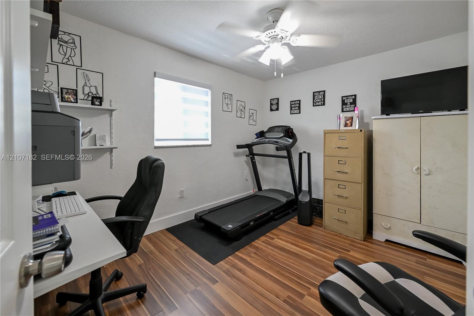 Room featuring wood-finish flooring, a ceiling fan with integrated lighting, a wall-mounted shelving unit, and a window with a roller shade