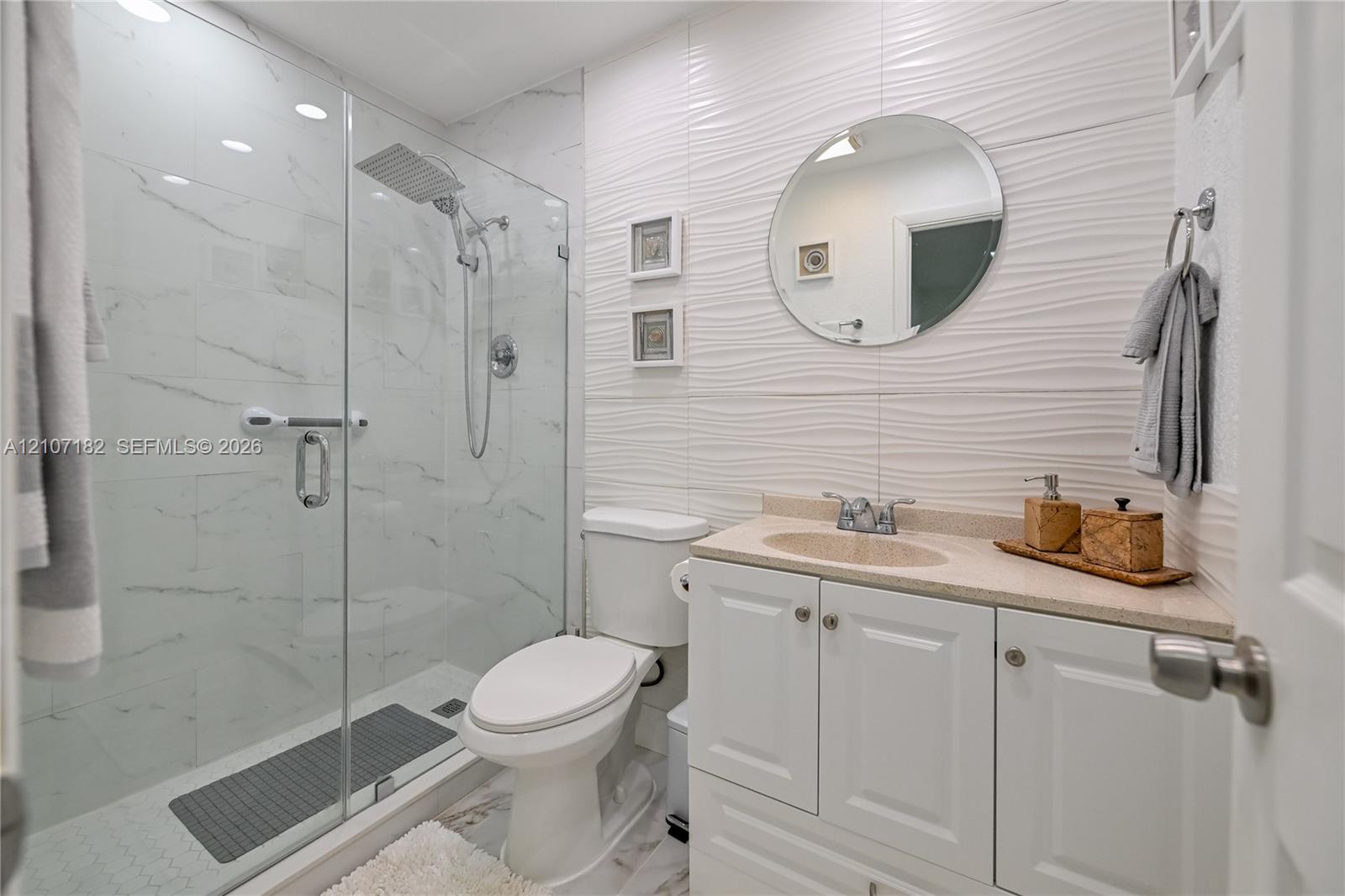 Bathroom featuring a glass-enclosed shower with rainfall showerhead, white vanity with integrated basin, stone-surface countertop, and textured wall tile