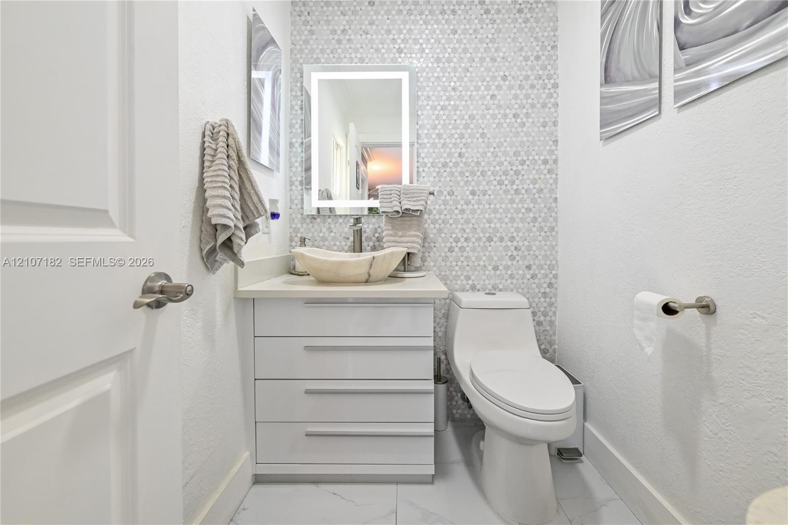 Bathroom featuring a stone vessel sink, white vanity with integrated cabinetry, mosaic tile wall accent, and illuminated vanity mirror