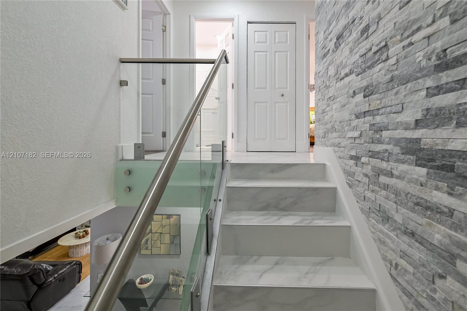 Staircase featuring white stone-finish treads, a stacked-stone accent wall, and a glass panel railing with a metallic handrail