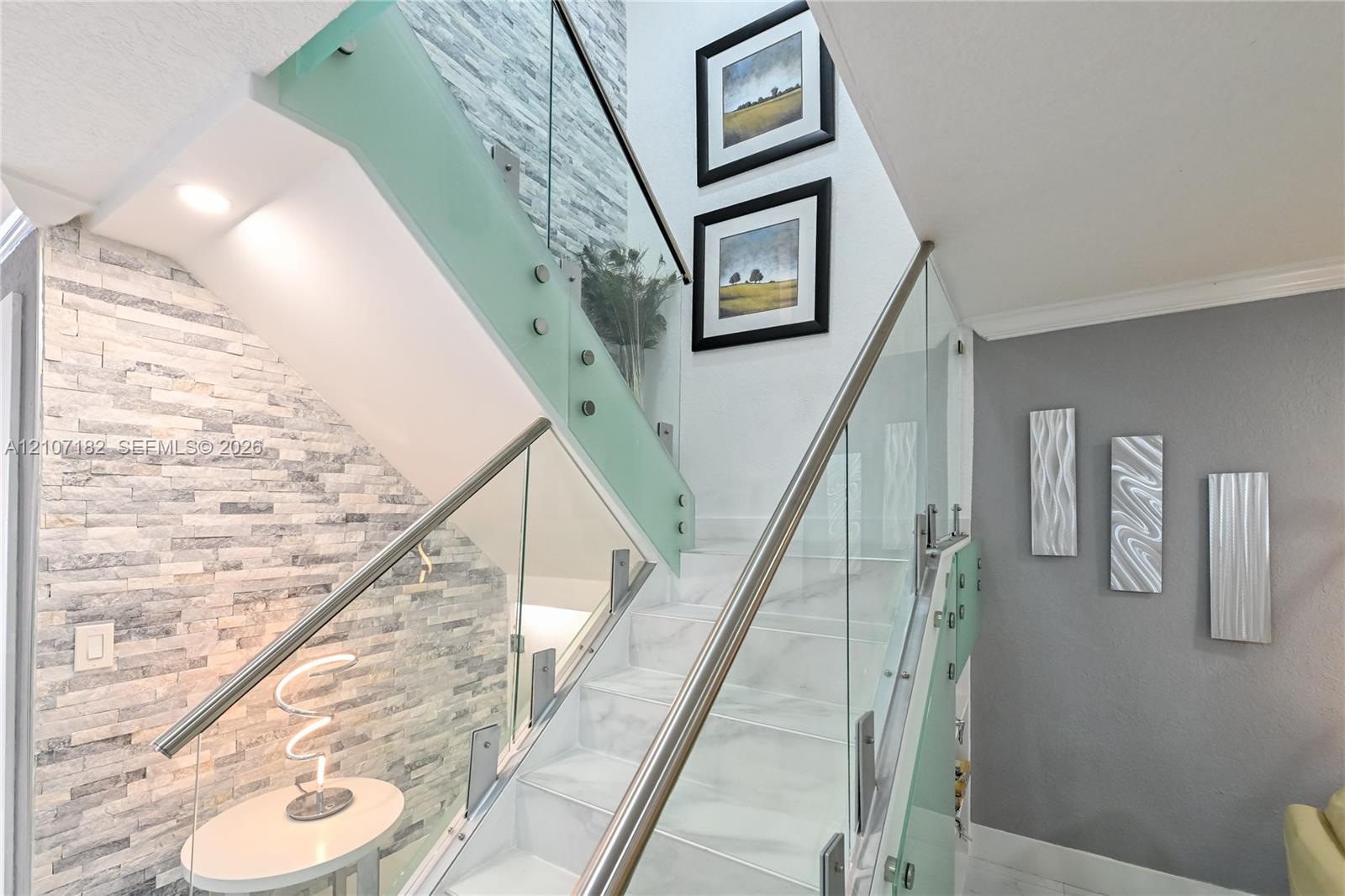 Staircase featuring white stone-finish treads, glass panel railing with stainless steel hardware, and an accent wall composed of stacked ledger stone