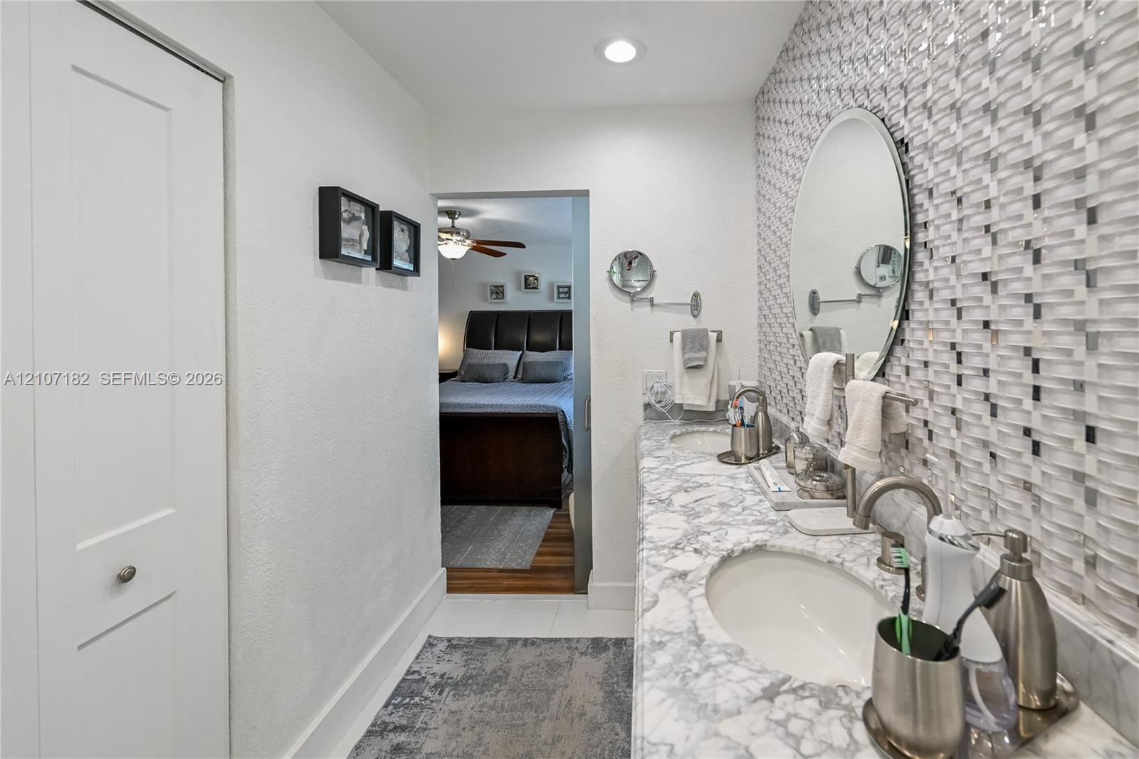 Dual-sink vanity with patterned stone countertop, custom mosaic tile backsplash, recessed lighting, and oval wall-mounted mirrors