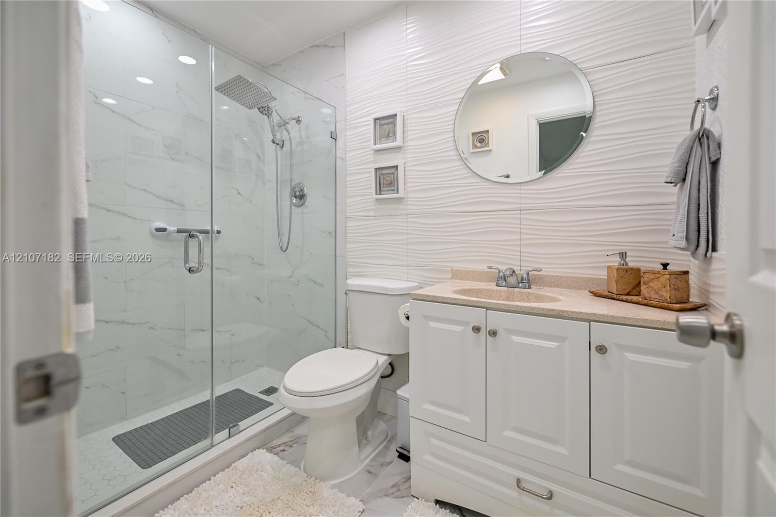 Walk-in shower with floor-to-ceiling stone-patterned tile and rainfall showerhead, white vanity with integrated countertop, and textured accent wall tiling