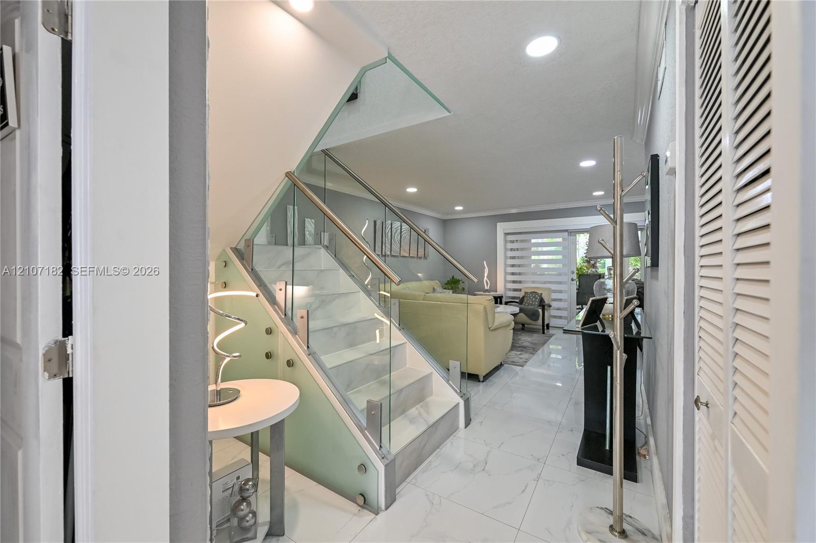 Staircase featuring white stone-finish steps, glass balustrade, and stainless steel handrail, complemented by large-format white tile flooring and recessed ceiling lighting