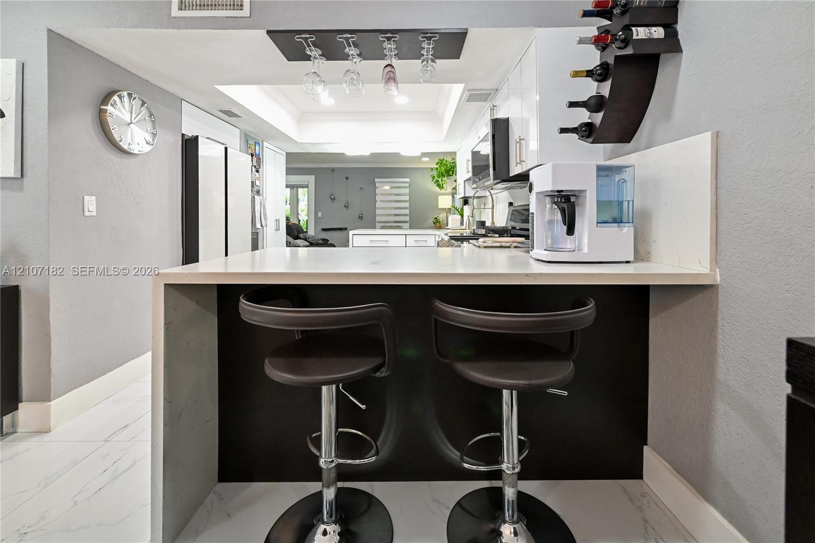 Waterfall peninsula featuring a solid-surface countertop and contrasting dark cabinetry, overhead wine glass rack, tray ceiling, and white floor tiling with veining
