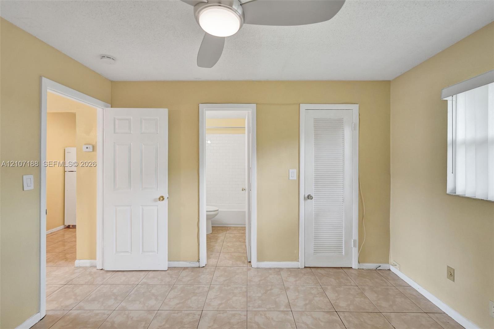 Neutral-toned room featuring light-colored tile flooring, a central ceiling fan with an integrated light fixture, a louvered-panel closet door, and interior paneled doors
