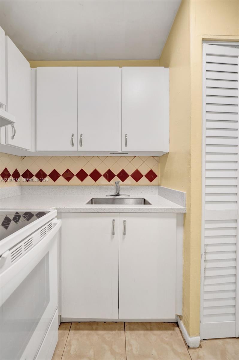 Kitchenette featuring white cabinetry, a neutral solid-surface countertop, stainless steel sink with a chrome faucet, diamond-patterned accent tile backsplash, and louvered closet doors