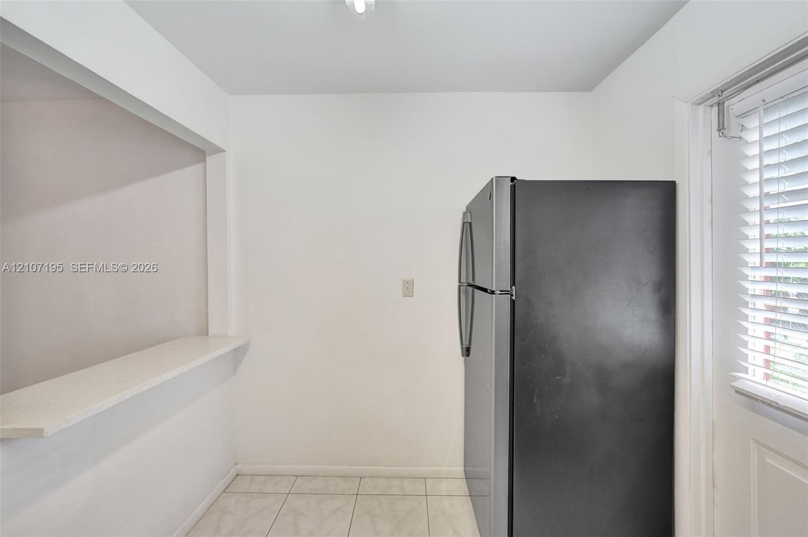 Built-in pass-through ledge, stainless steel refrigerator, white tile flooring, and window with fitted horizontal blinds