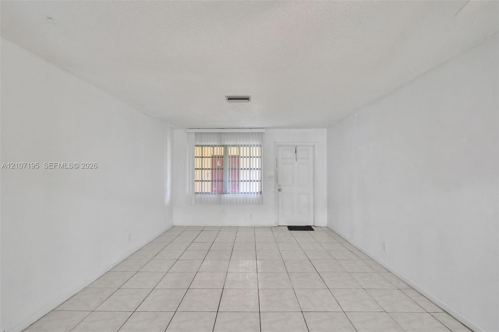 Spacious room featuring neutral tile flooring, smooth white walls, vertical window blinds, and a paneled entry door