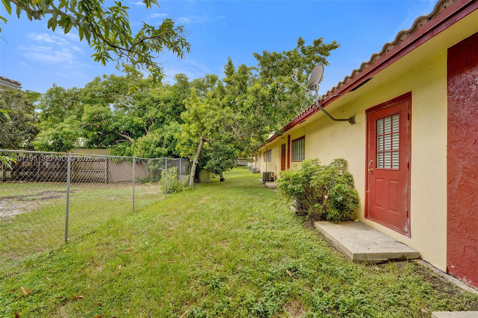 Exterior side yard featuring a chain-link perimeter fence, mature trees, concrete door step, and a red door with integrated window panels