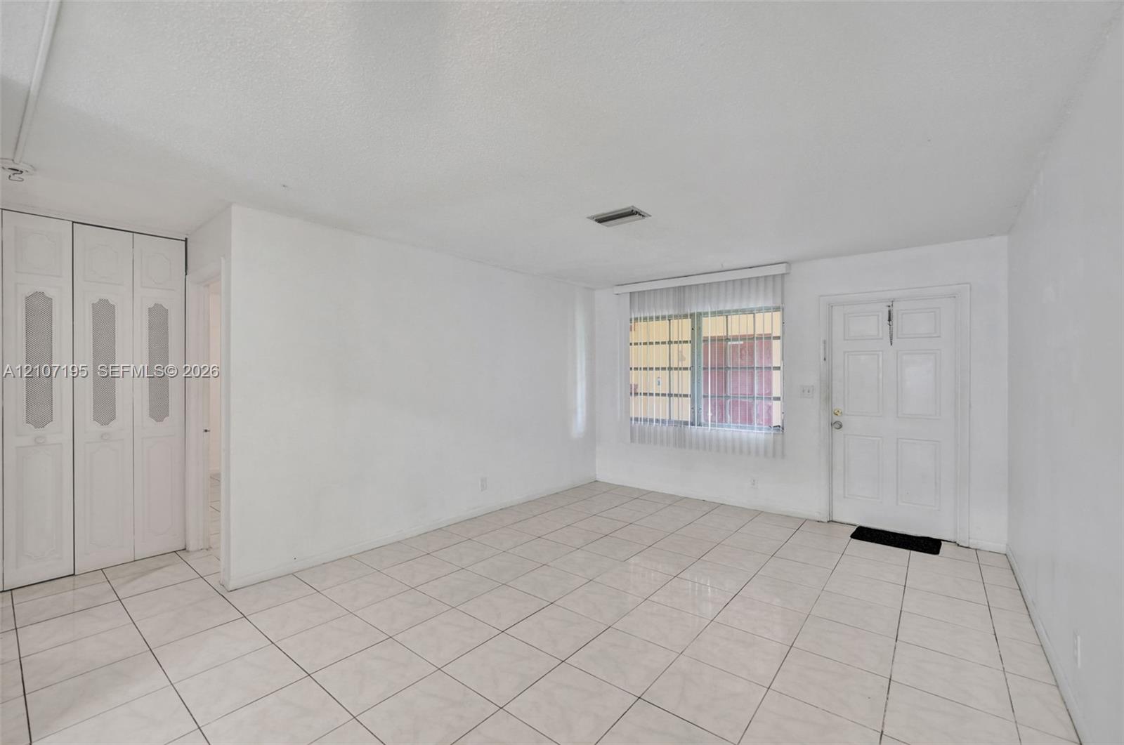 Living area featuring light-toned tile flooring, built-in floor-to-ceiling closet cabinetry, a multi-pane window with vertical blinds, and a paneled entry door