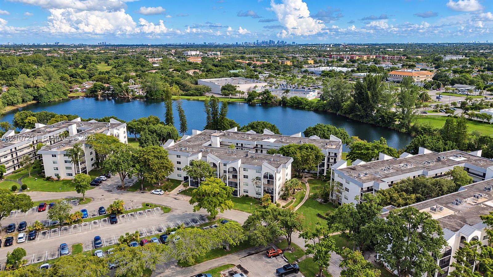 Aerial perspective of a multi-story residential complex featuring arched architectural details, private balconies, paved surface parking, and extensive waterfront grounds with mature landscaping