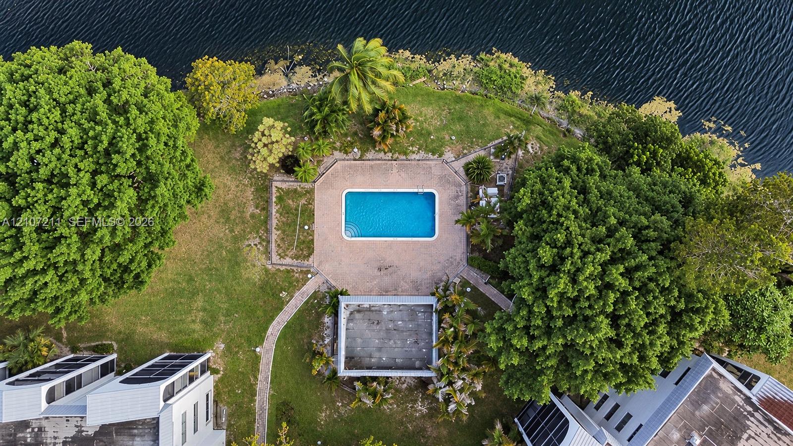 Aerial perspective of a waterfront property featuring a rectangular pool, paved deck, mature trees, and a manicured lawn