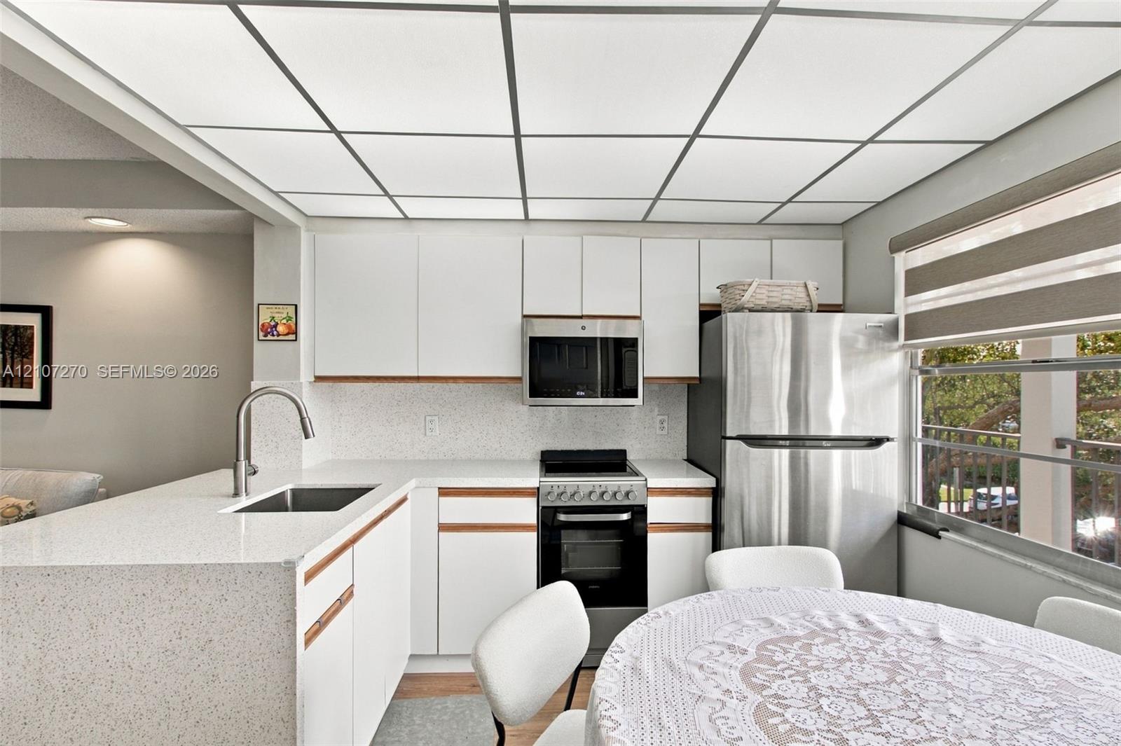 Kitchen featuring white flat-panel cabinetry, light-toned solid surface countertops, a stainless steel undermount sink with a gooseneck faucet, built-in stainless steel appliances, and a grid-style drop ceiling