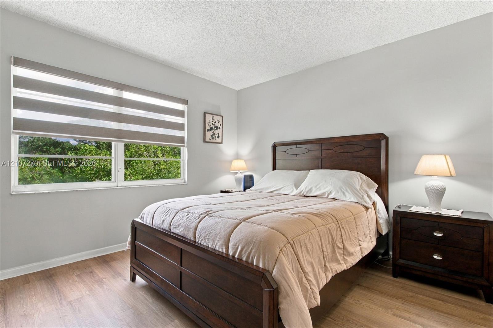 Bedroom featuring wood-finish flooring, textured ceiling, and large window with adjustable zebra-pattern roller shades