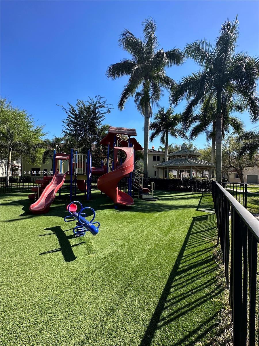 Outdoor recreational space featuring a play structure with dual slides, a spring rider, an octagonal gazebo, mature palm trees, and synthetic turf enclosed by metal fencing
