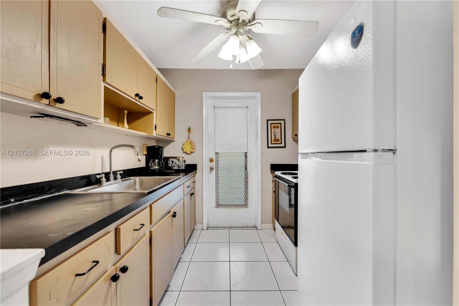 Kitchen featuring cabinetry with contrasting dark countertops, integrated stainless steel sink with goose-neck faucet, white tile flooring, and a ceiling-mounted light fixture with fan