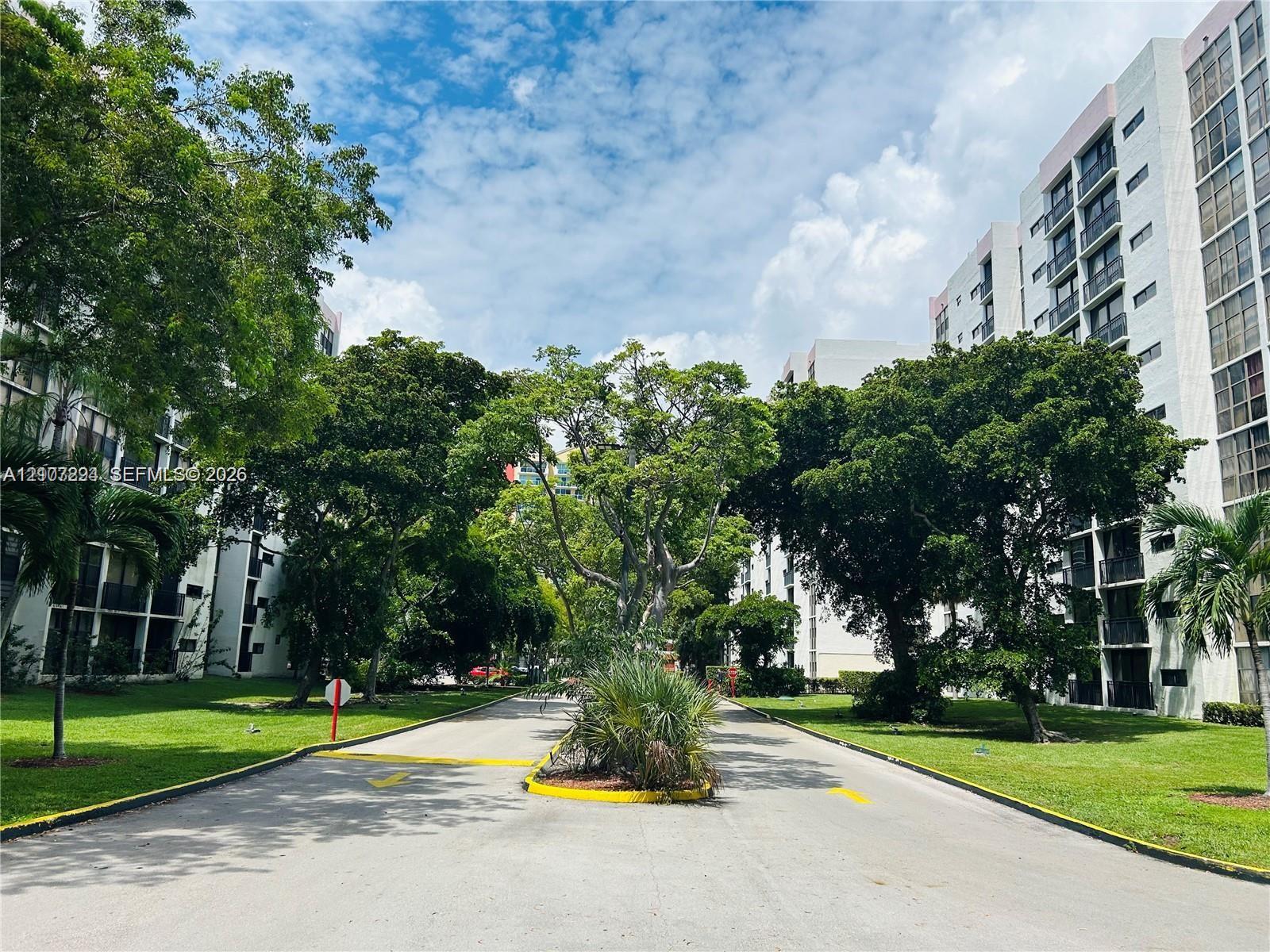 Multi-story residential complex featuring white stucco facades, private balconies, expansive green space, mature canopy trees, and paved thoroughfares