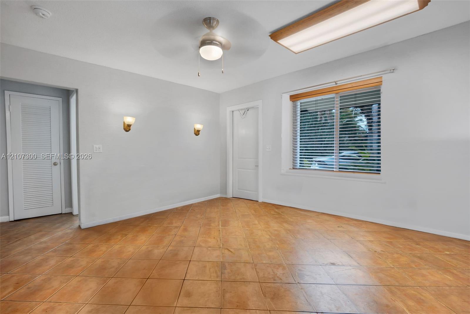 Interior space featuring tile flooring, dual wall-mounted light fixtures, a ceiling fan with integrated light, a louvered door, and a recessed window with horizontal blinds