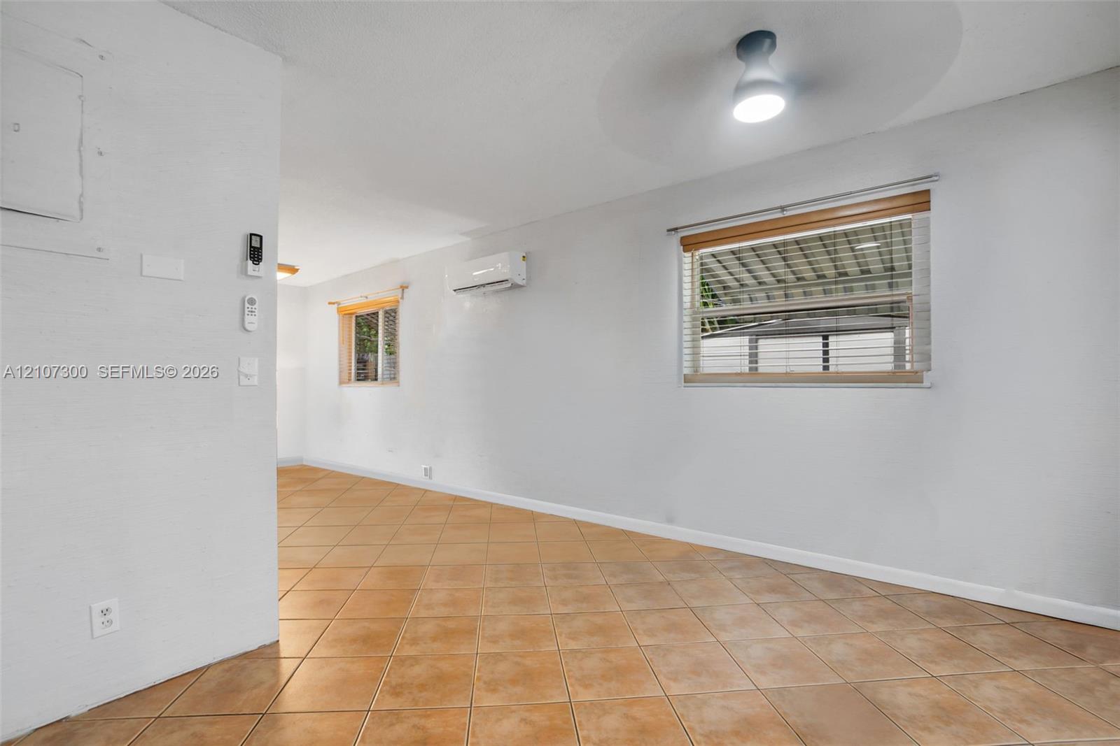 Neutral-toned interior space featuring ceramic tile flooring, wall-mounted ductless air conditioning unit, ceiling-mounted lighting, and horizontal window blinds