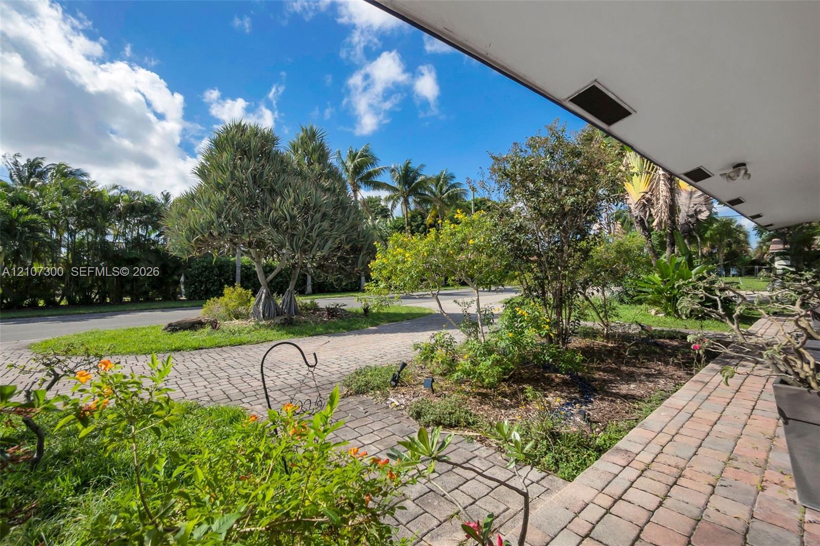 Covered outdoor porch with stone-paver flooring, extensive mature landscaping, and multi-patterned stone-paver driveway