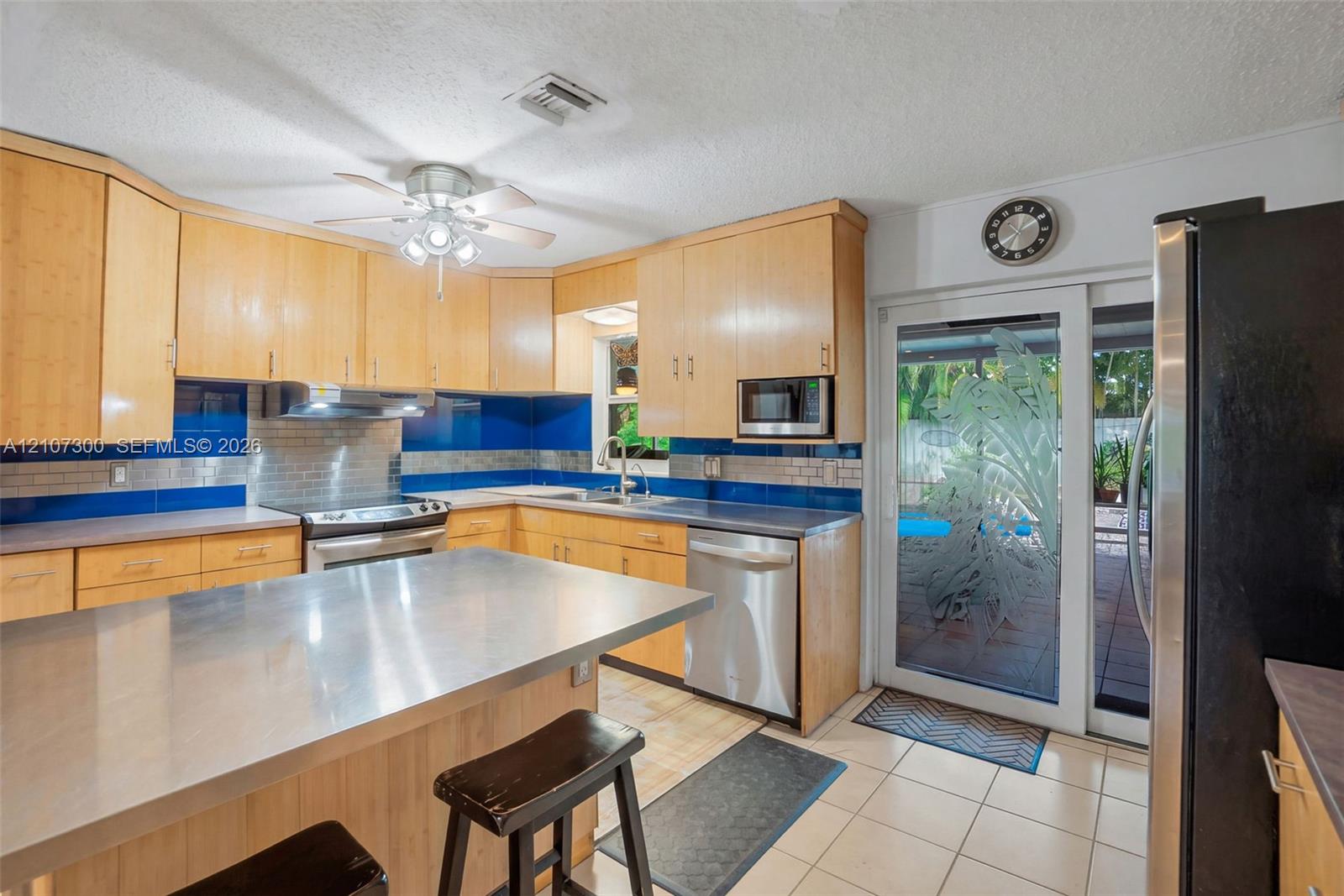 Kitchen featuring light wood cabinetry, stainless steel island countertop, tile flooring, glass sliding door with etched detailing, and integrated stainless steel appliances