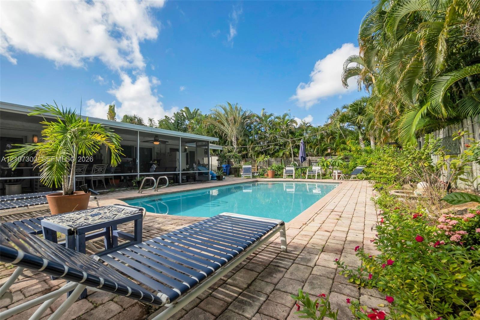 Outdoor area featuring a rectangular in-ground swimming pool, expansive paver patio, and perimeter privacy fencing surrounded by mature palm trees
