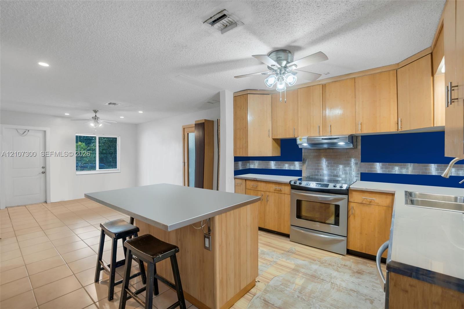 Kitchen featuring wood cabinetry, a central island with a solid-surface countertop, stainless steel range with overhead vent hood, and tiled flooring