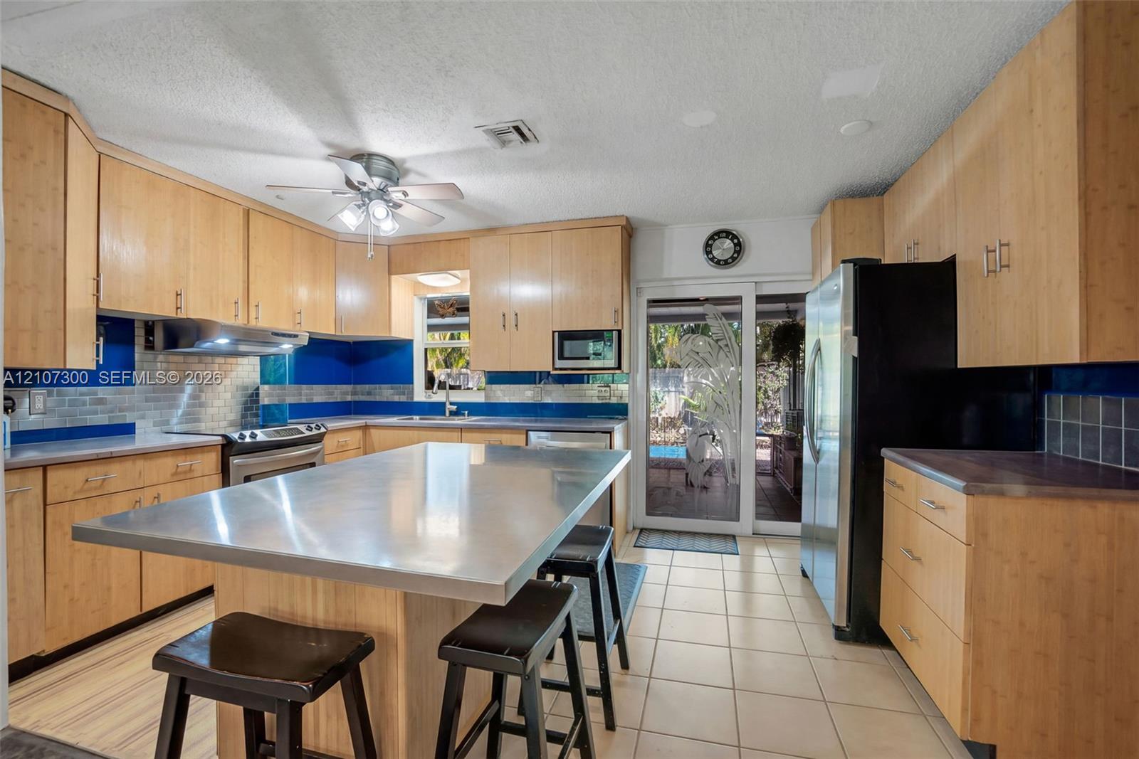Kitchen featuring light wood-finish cabinetry, a stainless steel island countertop, tile flooring, blue glass tile backsplash, and stainless steel appliances