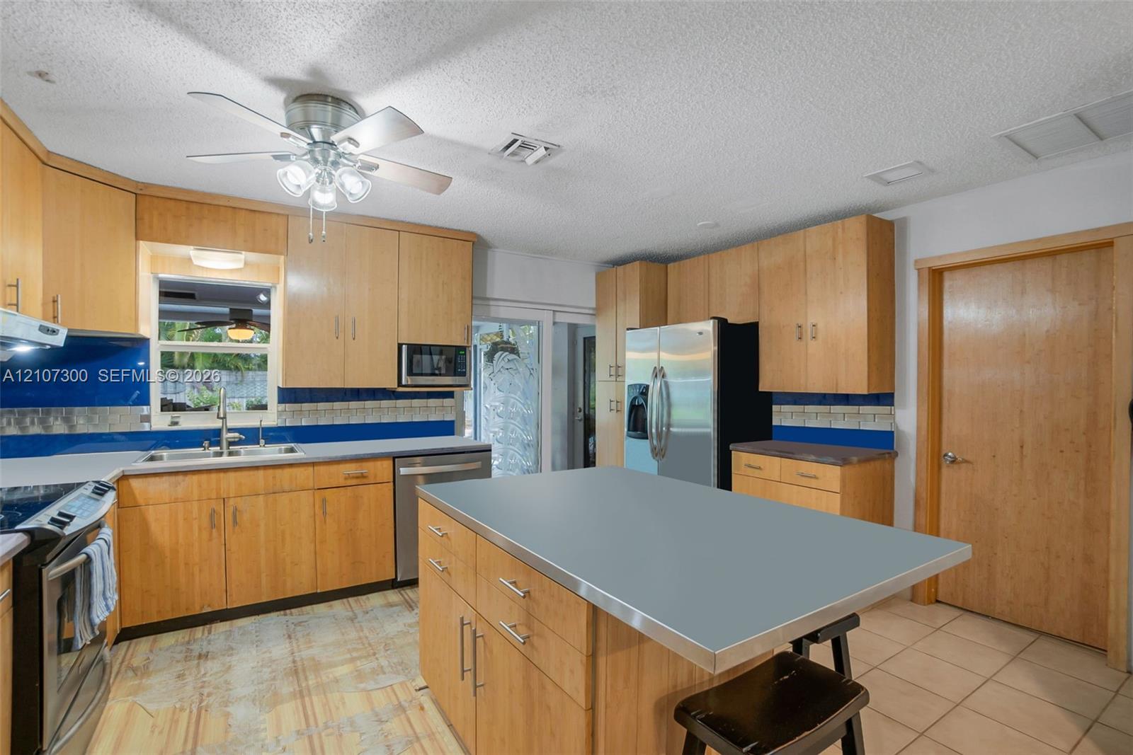 Kitchen featuring light-finish cabinetry, a central island with a solid-surface countertop, stainless steel appliances, blue tiled backsplash, and tile flooring
