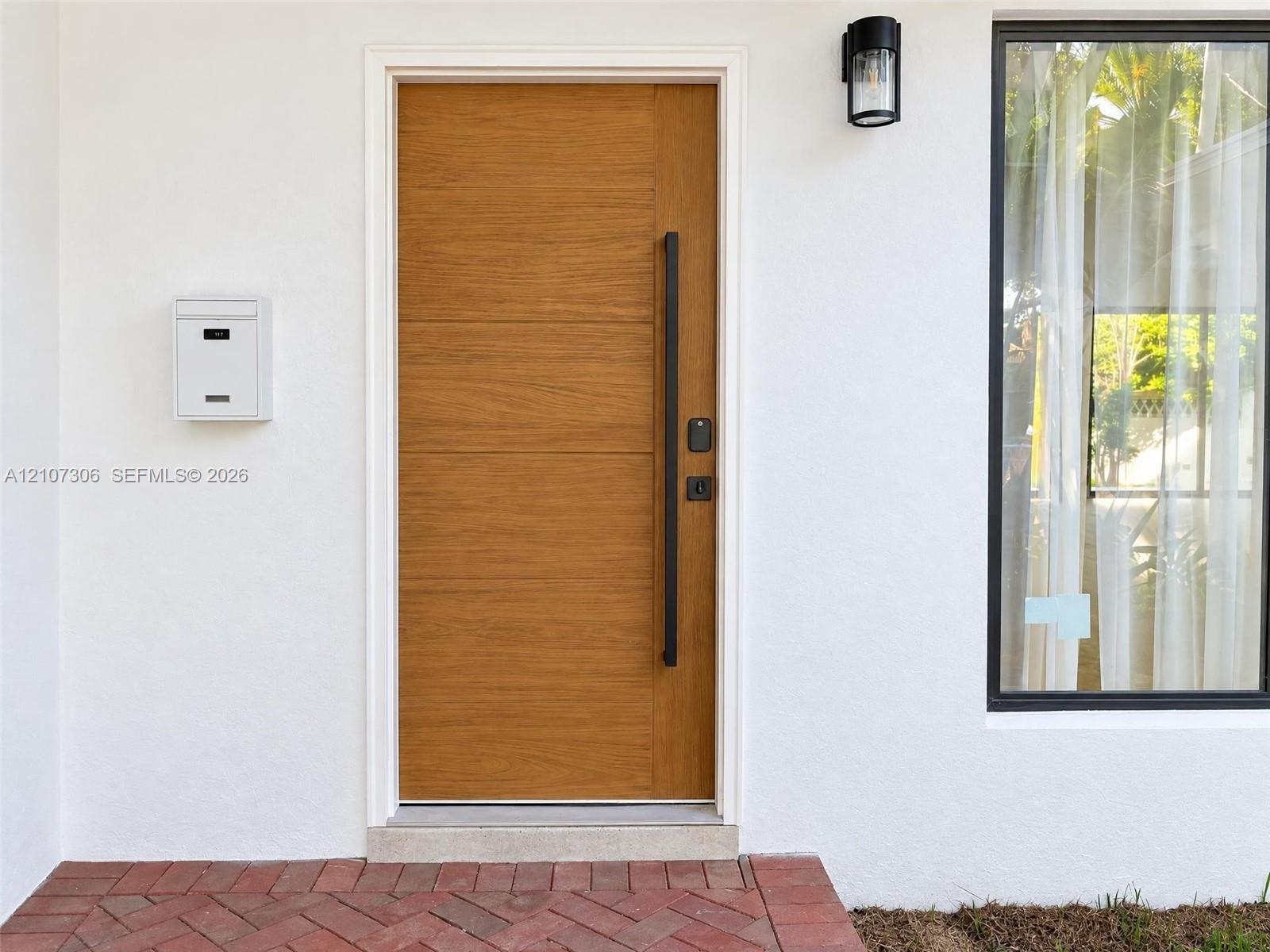 Wood-finish front door with a vertical matte black pull handle, white stucco exterior, exterior wall-mounted lighting, and herringbone-patterned brick entryway