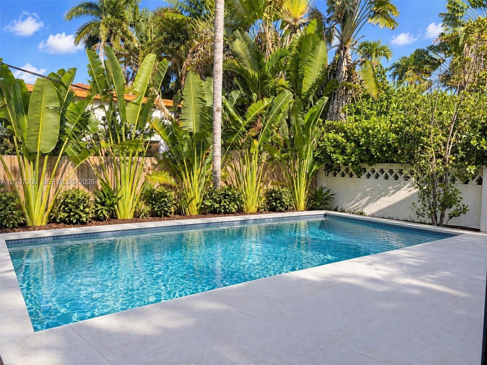 In-ground swimming pool with mosaic tile border, light-toned stone-finish patio, decorative masonry screen wall, and perimeter tropical landscaping