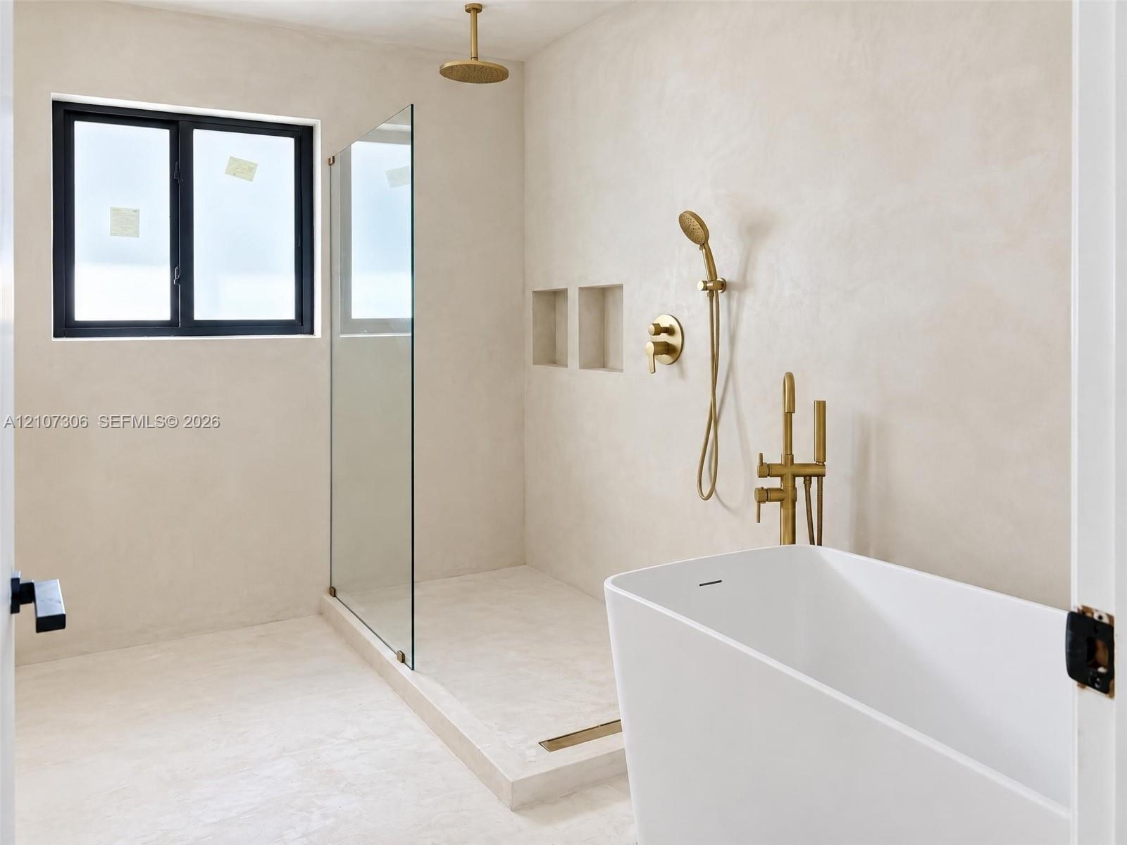 Freestanding soaking tub with brass fixtures, glass-enclosed walk-in shower, dual recessed wall niches, and matte-finish plaster wall treatment