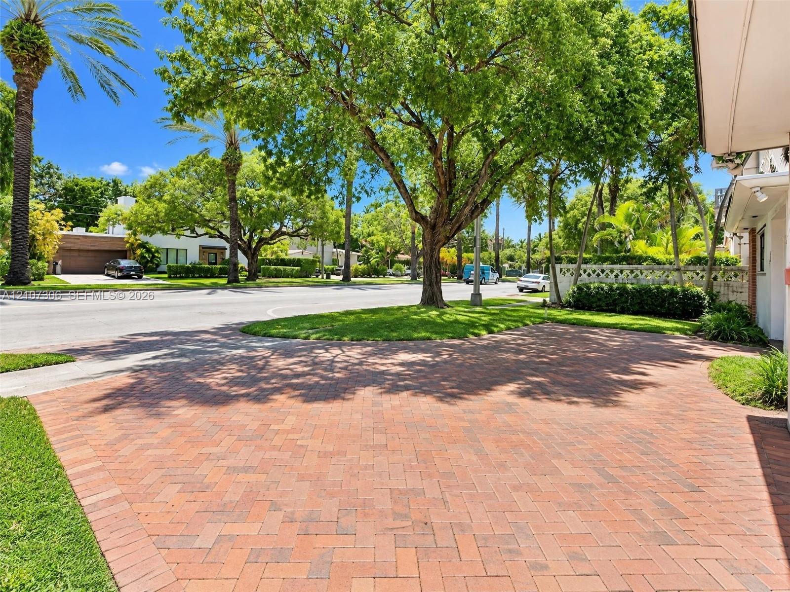 Brick-paver driveway with a herringbone pattern, mature trees, manicured lawn, and perimeter landscaping
