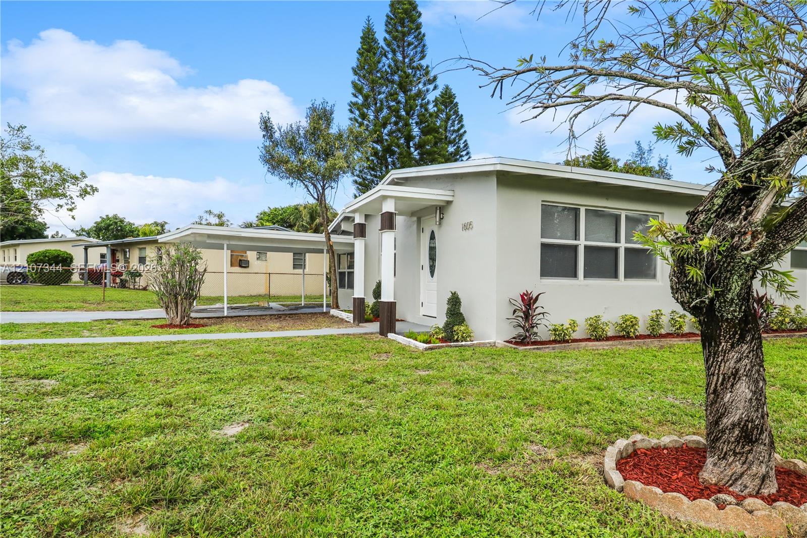 Single-story exterior featuring a covered carport, concrete walkway, and decorative masonry tree surround