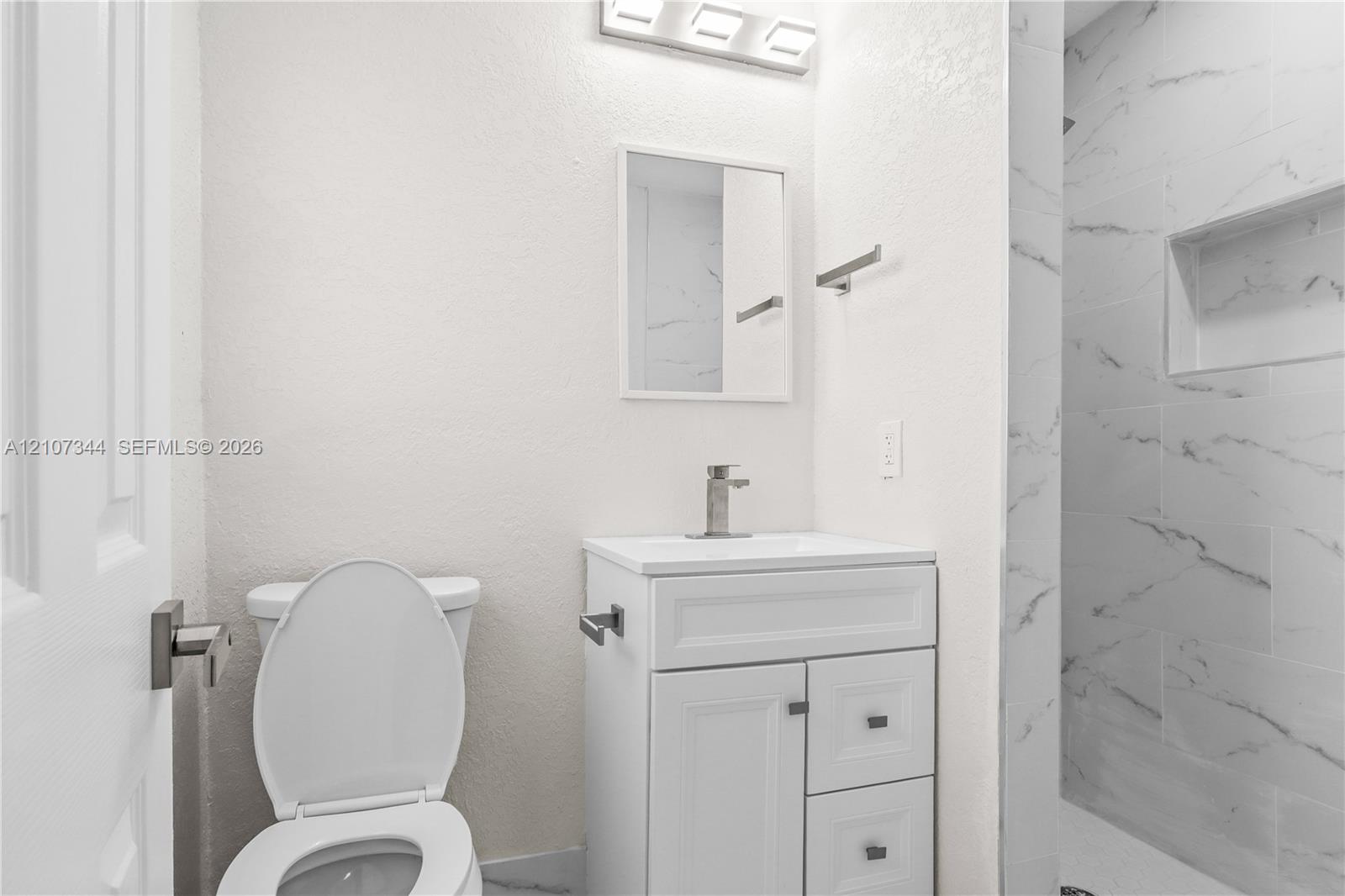 White vanity with integrated basin, polished metal faucet, wall-mounted three-light fixture, and shower enclosure featuring large-format tile with integrated recessed niche
