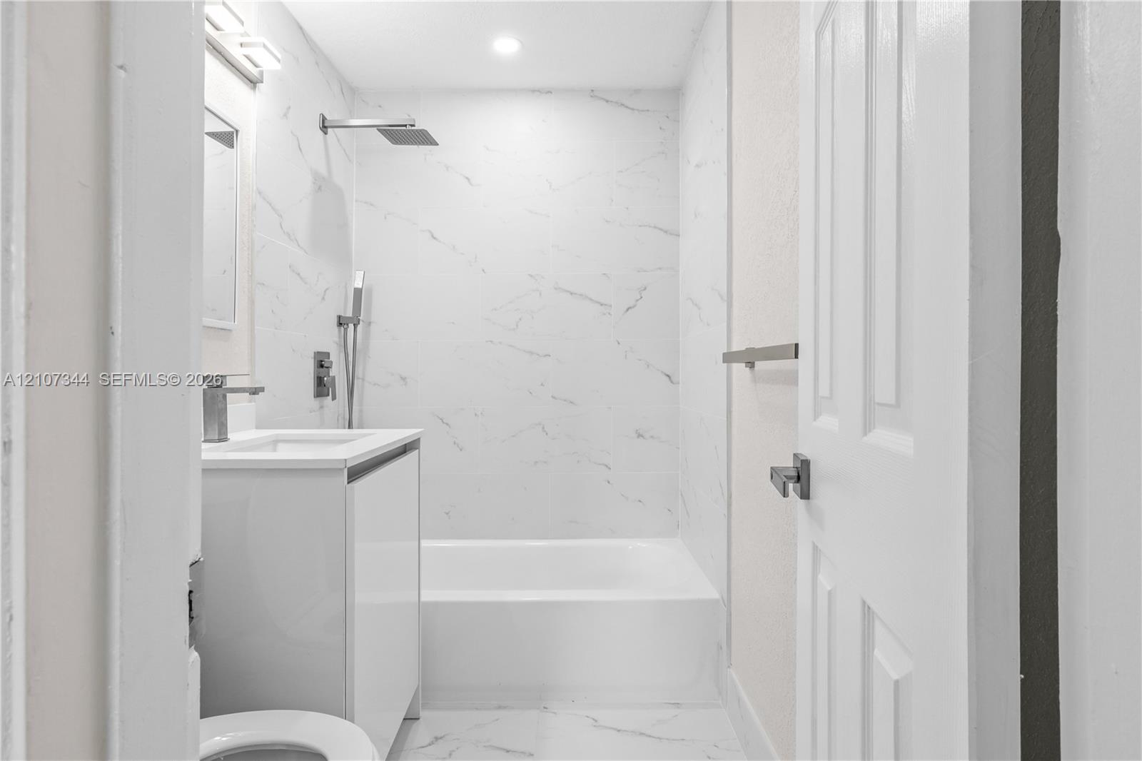 Full-height stone-finish wall tile, white vanity cabinet, rectangular undermount sink, rainfall showerhead with handheld sprayer, and recessed lighting