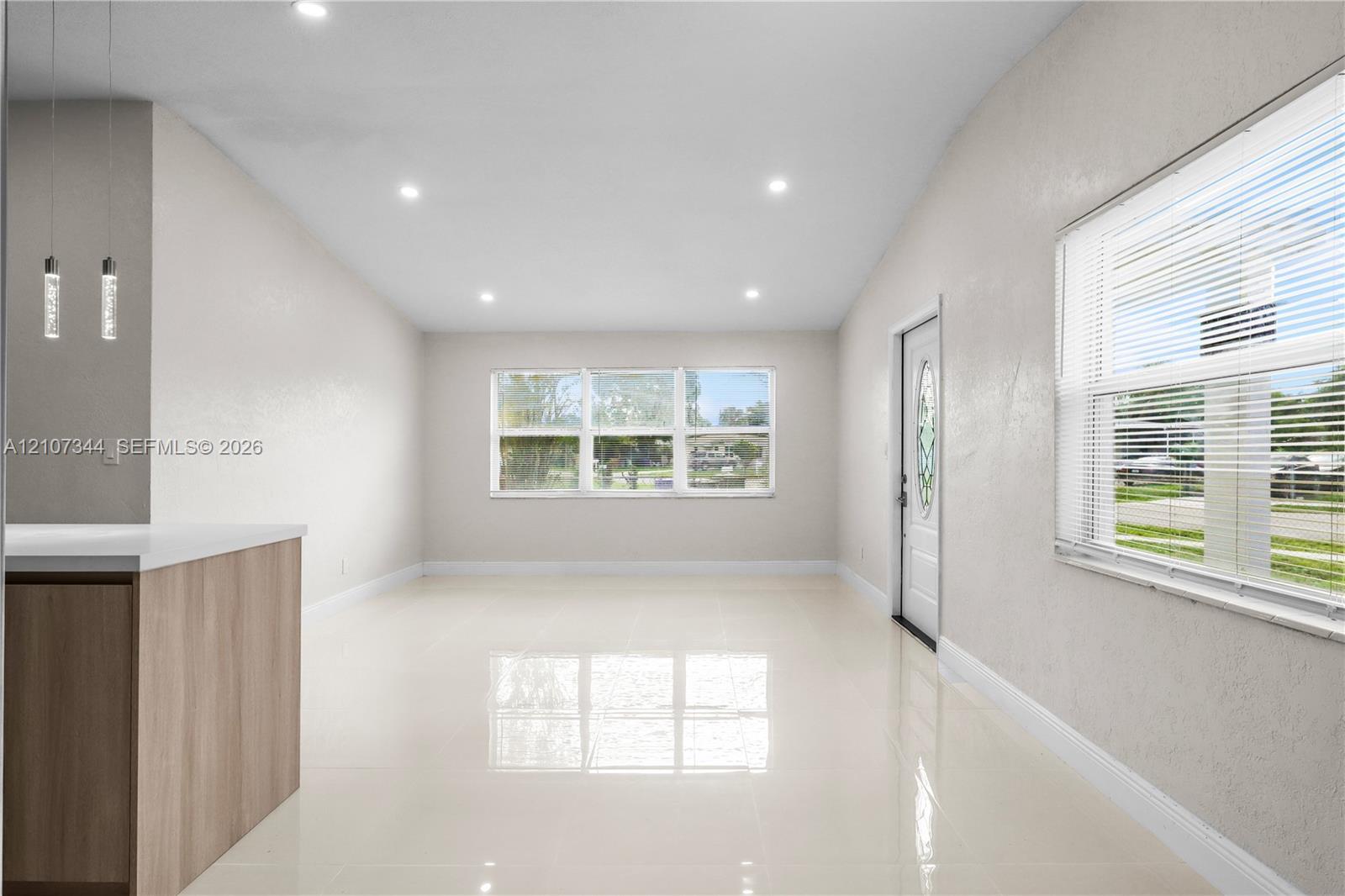 Open-concept room featuring light-toned tile flooring, recessed ceiling lighting, a wood-finish peninsula with a solid-surface countertop, and dual pendant light fixtures