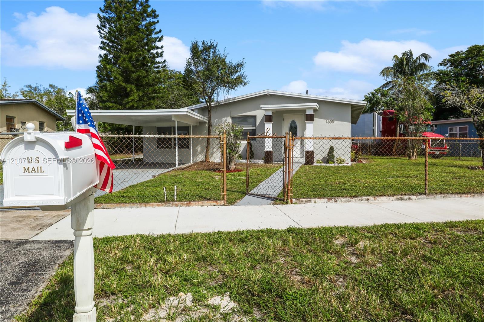 Single-story exterior featuring a covered carport, concrete walkway, and chain-link perimeter fencing