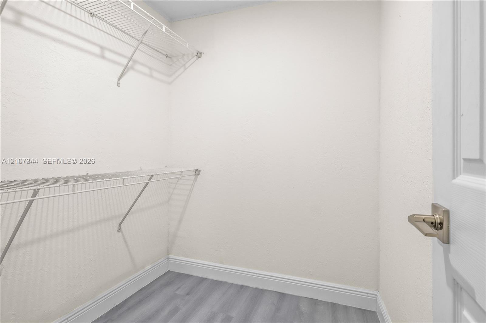Storage space featuring dual-level wire shelving and wood-finish flooring, accessed by a paneled door with metallic lever hardware