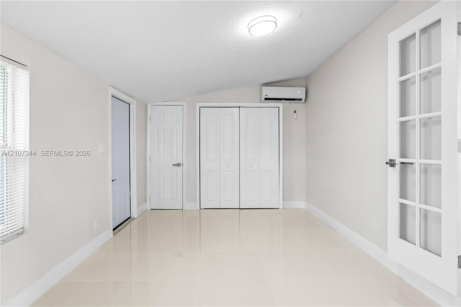 Neutral-toned interior featuring large-format tile flooring, double-door closet cabinetry, wall-mounted ductless climate control unit, and a French-style door with glass paneling