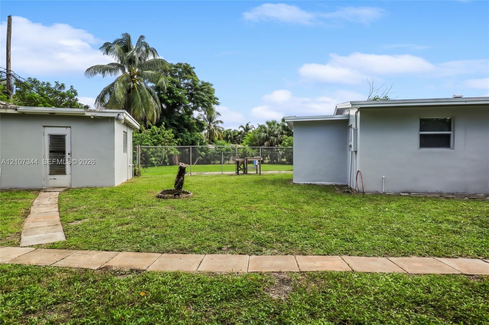 Expansive backyard featuring a manicured lawn, concrete paver pathway, chain-link perimeter fencing, and mature palm trees