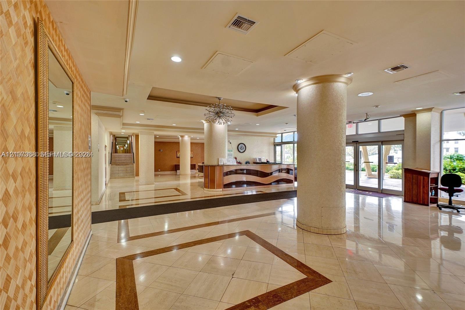 Lobby featuring polished stone flooring with inlaid borders, structural columns, recessed lighting, a built-in reception desk, and a decorative chandelier